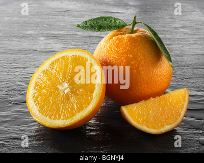 Ganze und geschnittene frische Orangen mit Blättern vor schwarzem Hintergrund Stockfoto