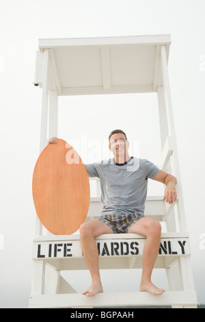 Reifer Mann sitzt auf einer Rettungsschwimmer-Hütte und hält ein Surfbrett Stockfoto