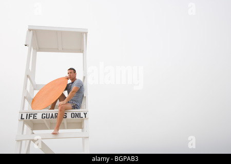 Niedrigen Winkel Blick auf ein reifer Mann sitzt auf einer Rettungsschwimmer-Hütte und hält ein Surfbrett Stockfoto