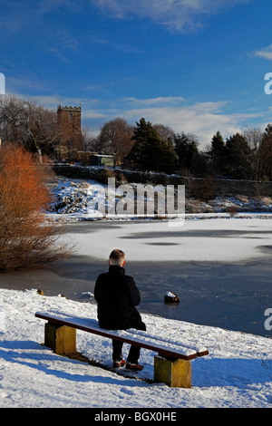 Mann sitzt in der Sonne auf Schnee bedeckten Bank Blick über gefrorene Duddingston Loch mit Kirk Hintergrund Edinburgh Schottland Stockfoto