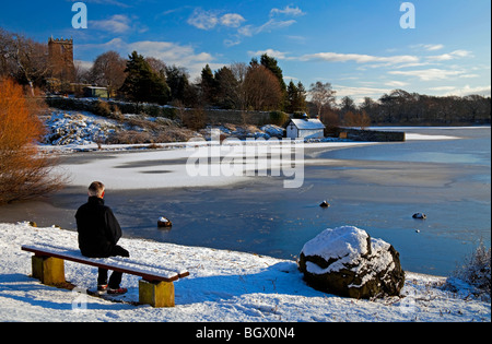 Mann sitzt in der Sonne auf Schnee bedeckten Bank Blick über gefrorene Duddingston Loch mit Kirk Hintergrund Edinburgh Schottland Stockfoto