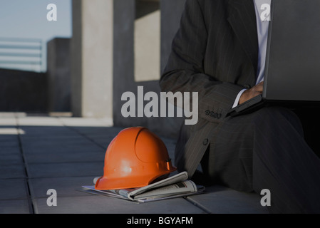 Mann sitzt auf dem Bürgersteig mit Laptop, orange Schutzhelm auf Seite vor Zeitung eingestellt Stockfoto