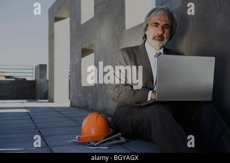 Mann sitzt auf dem Bürgersteig mit Laptop, Schutzhelm seitlich unfertigen Gebäude im Hintergrund festlegen Stockfoto