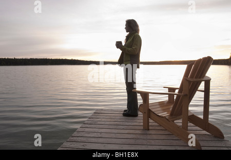 Frau stehend auf Dock hält Tasse Kaffee bei Sonnenuntergang, Clear Lake, Manitoba Stockfoto