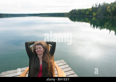 Frau mit geschlossenen Augen sitzend im Liegestuhl am dock neben Clear Lake, Riding Mountain National Park, Manitoba, Kanada Stockfoto