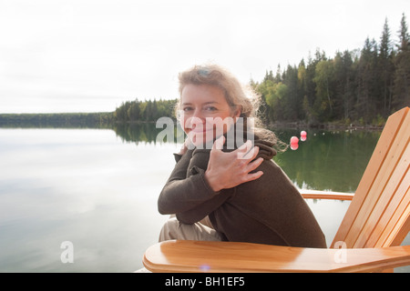 Lächelnde Frau wickelt Arme um selbst sitzt im Liegestuhl an Deck neben Clear Lake, Manitoba, Kanada Stockfoto