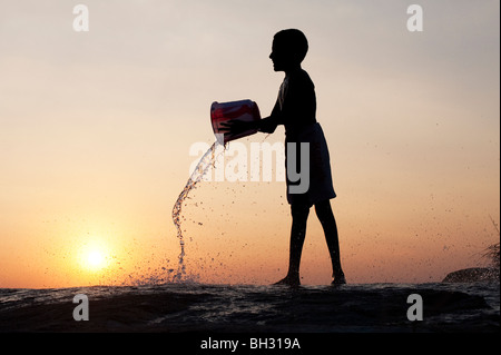 Indischer junge gießt Wasser auf einem Felsen aus einem Eimer bei Sonnenuntergang. Andhra Pradesh, Indien Stockfoto