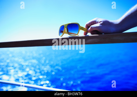 Die Hand eines Mannes hält sich an ein paar Retro-gelbe Sonnenbrille ruht auf einem Handlauf aus Holz auf einem Kreuzfahrtschiff gegen ein blaues Meer Stockfoto