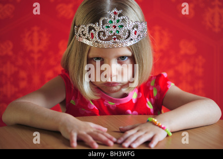 Junges Mädchen mit einem roten Kleid und eine Prinzessin Krone sitzt an einem Tisch, runzelt die Stirn mit einem miserablen Ausdruck Stockfoto