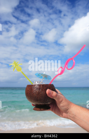 Eines Mannes Hand mit einem tropischen cocktail in einer Kokosnussschale auf einem einsamen tropischen Strand Stockfoto