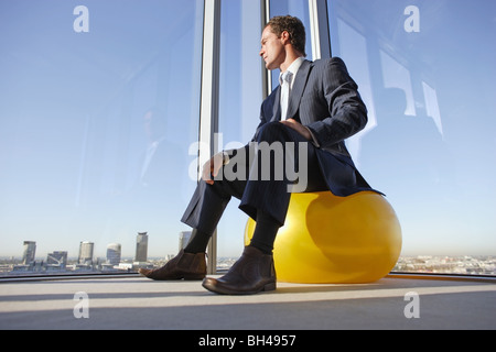 Ein Geschäftsmann sitzt auf einem Swiss-Ball in der Ecke eines Büros, Blick aus dem Fenster über die Wolkenkratzer Stockfoto