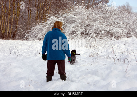 Dame in blauem Vlies spielt im Schnee, während ihr tief verschneiten Labradoodle Welpen beobachtet und wartet, dass ihr darauf Stockfoto