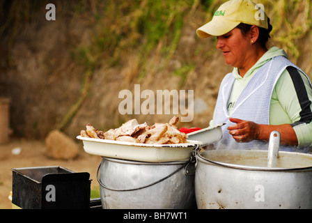 Ecuador, Otavalo, Seitenansicht einer Frau mit einer leichten blau karierte Schürze und eine gelbe Kappe Kochen in einem Stall mit tiefen gebratenen c Stockfoto