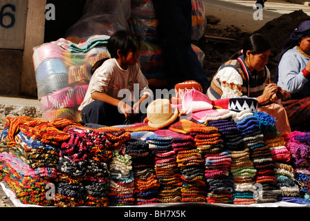 Ecuador, Otavalo, junge Mädchen sitzen durch Stapel von bunten wollene Kleidung und Hüte auf dem Display in Straßenmarkt Stockfoto