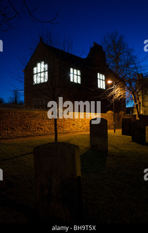 Gruseligen alten Schulhaus mit beleuchtet Windows in der Abenddämmerung mit Grabsteinen im Vordergrund Stockfoto