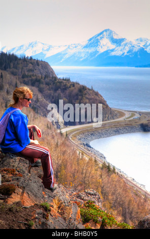 Frau sitzt auf Bergrücken mit Blick auf Turnagain Arm mit Seward Highway und Chugach Mountains im Hintergrund/n Stockfoto