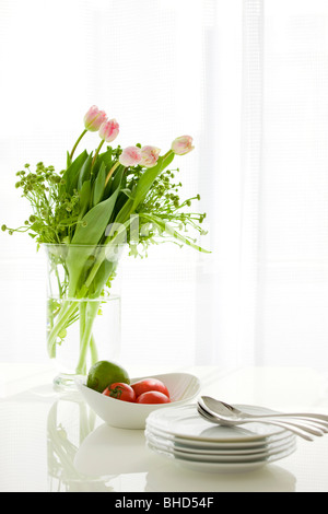 Vase mit Lilien auf Tabelle mit Tomaten, Äpfel, Teller und Löffel Stockfoto