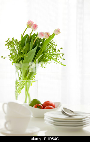 Vase mit Lilien auf Tabelle mit Tomaten, Äpfel, Teller und Löffel Stockfoto