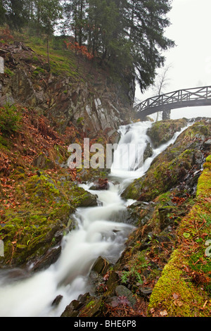 Ruhiger Wasserfall, der unter einer hölzernen Brücke in einem üppigen Park der Tschechischen Republik fließt, mit Stufen, die auf einen grasbewachsenen Hügel führen Stockfoto