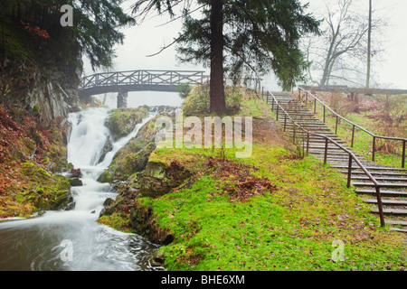 Ruhiger Wasserfall, der unter einer hölzernen Brücke in einem üppigen Park der Tschechischen Republik fließt, mit Stufen, die auf einen grasbewachsenen Hügel führen Stockfoto