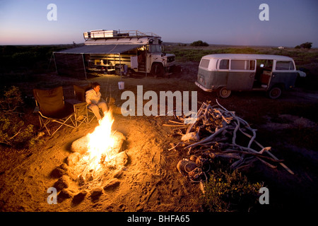 Frau sitzt am Lagerfeuer, einen amerikanischen Schulbus und einem alten VW-Bus im Hintergrund, Conejo, Baja California Sur, Mexiko Stockfoto