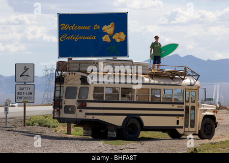 Eine 18 Jahre alte Teenager mit einem Surfbrett stehen am Anfang einer amerikanischen Schulbus an der California welcome Schild, Interstate 15, Stockfoto