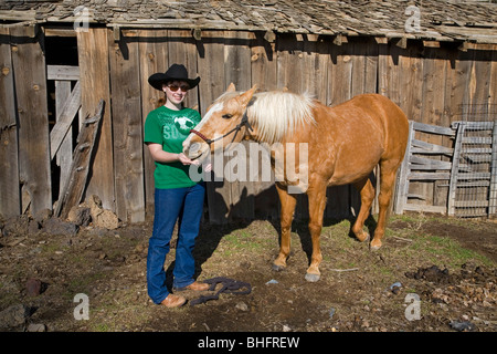 Ein junges Cowgirl mit einem zehn Gallonen-Hut und ihrem Palomino-Pferd vor einer alten Scheune. Stockfoto