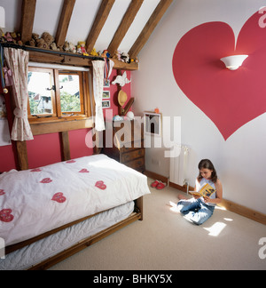 Kleines Mädchen im Land-Ferienhaus Kinderzimmer mit roten Herzen gemalt auf Wand und Herz-Motiv Bettdecke auf Bett Stockfoto