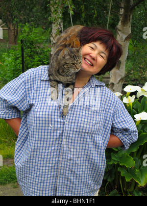 Gesunde reife Eurasierin (Chinesisch/Italienisch) in ihrem Garten mit einer Katze auf der Schulter Stockfoto