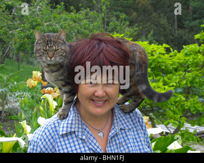Gesunde reife Eurasierin (Chinesisch/Italienisch) in ihrem Garten mit einer Katze auf der Schulter Stockfoto