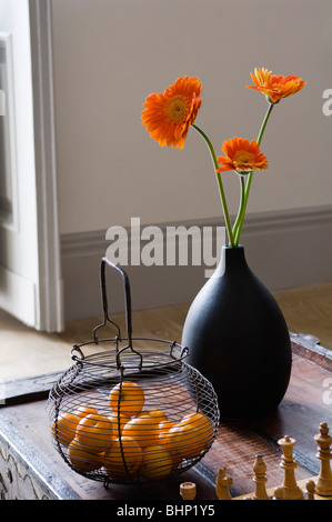 Orange Gerbera Blumen in schwarzer Vase auf Tischplatte mit Metallkorb Stockfoto