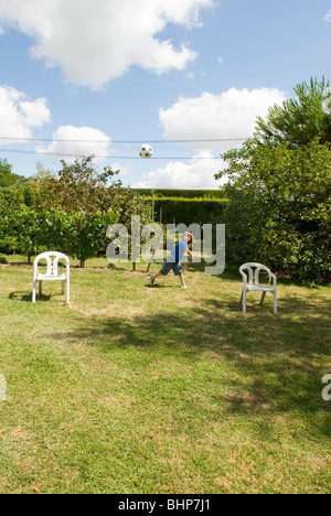 ein sieben Jahre alter Junge wirft einen Fußball in die Luft zwischen zwei provisorische Stühle als Torpfosten in einem Garten Stockfoto