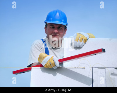 Mason tragen blaue Helm posiert auf weißer Blockwand hält Rotpegel über blauen Himmel ruht Stockfoto