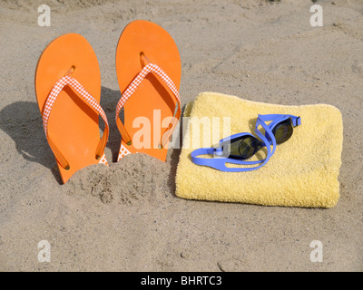 Orange Flip Flops, die vertikal in Strandsand und Schwimmbrille auf gelben Badehandtuch getrieben Stockfoto