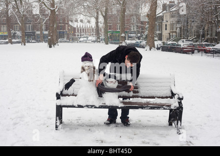Ein Mann beugt sich hinüber zu den letzten Schliff, zwei kleine Schneemänner, der jetzt eine Parkbank auf Goose Green in East Dulwich besetzen. Stockfoto