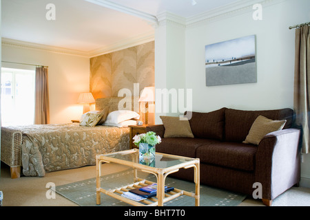 Zuckerrohr und Glas Tisch vor braunen Sofa im Stadthaus Schlafzimmer mit brennenden Lampen auf beiden Seiten des Bettes mit Beige Bettdecke Stockfoto