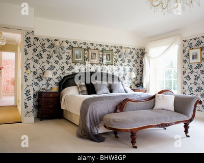 Graue Velours-Chaiselongue am Fuße des Bettes mit grauer Seide werfen im Schlafzimmer mit Schwarz + Weiß floral Tapete Stockfoto