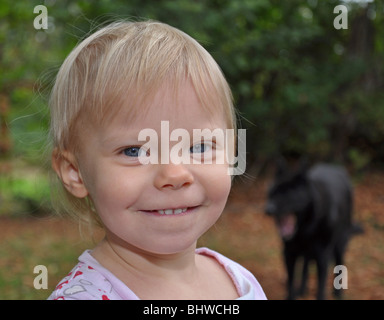 Dieses schöne blonden Haaren blauen Augen Kleinkind Mädchen ist in einer Nahaufnahme lächelnd und glücklich erschossen. Hintergrund mit Hund ist unscharf. Stockfoto