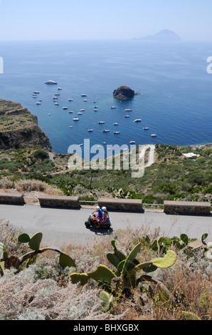 Ein paar Reiten auf einem Moped auf der Insel Salina, mit Blick auf ein Boot Bucht gefüllt, auf den Äolischen Inseln, Pollara, Sizilien, im Süden Italiens. Stockfoto