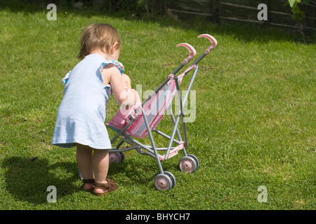 16 Monate altes Mädchen spielt mit Puppe und Buggy im Garten. Stockfoto