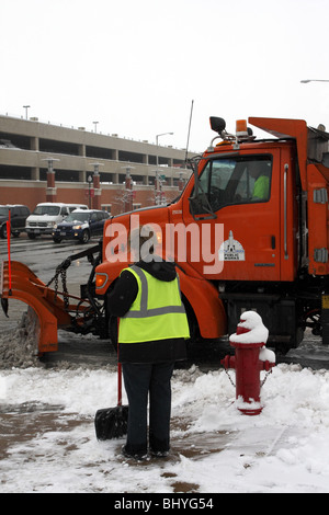 Frau mit Schneeschaufel Uhren aus Bürgersteig als kommunale LKW mit Schneepflug Klinge dreht sich eine Ecke. Stockfoto