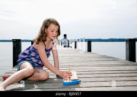 junge Mädchen spielen mit Spielzeugboot am Pier, während Vater im Hintergrund das Fischen Stockfoto