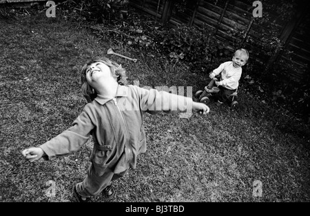 Ein drei Jahre altes Mädchen wirft den Kopf zurück mit Freude beim Spielen mit ihren jungen ein-Jahr-alte Bruder im Garten hinter dem Haus. Stockfoto