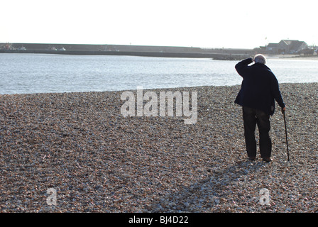 Alter Mann mit Stock zu Fuß am Strand von Lyme Regis. Cobb Hafen im Hintergrund Stockfoto
