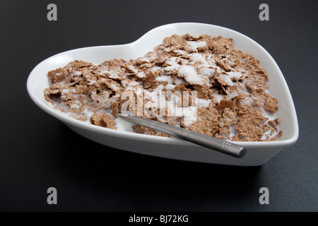 Herzförmige Müslischale oder Schale mit Löffel und Getreide auf schwarzem Hintergrund. Stockfoto