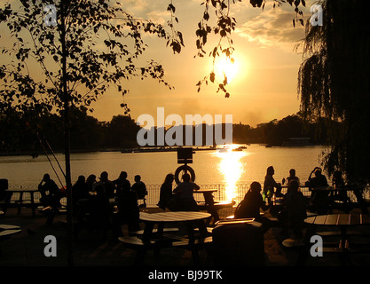 Sonnenuntergang über der Serpentine im Hyde Park, London mit Silhouetten von Menschen genießen Sie Getränke in entspannter Atmosphäre Stockfoto