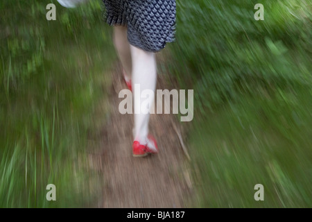 Mädchen mit blass weiße Beine, Sommerkleid und roten Schuhen laufen entlang eines Pfads, umgeben von grünen Rasen Stockfoto