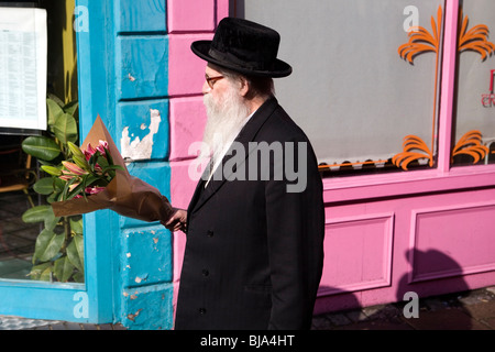 Senior orthodoxen jüdischen Mann mit schwarzem Hut und Mantel, rosa Lilien Blumen vorbei an bunten rosa und blauen Restaurant front Stockfoto