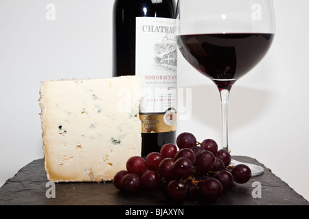 Stillleben mit Rotwein, einen Keil von Blauschimmelkäse, Trauben und eine Flasche Rotwein auf schwarzem Schiefer gegen einen weißen Hintergrund Stockfoto