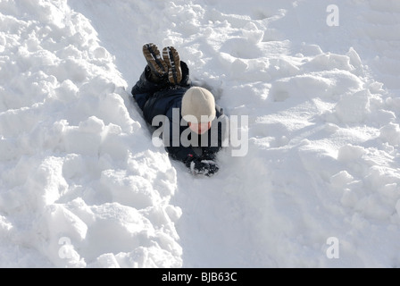 Lachendes Kind junge rutscht ein Schnee bedeckt Hill und spielen im Schnee Stockfoto